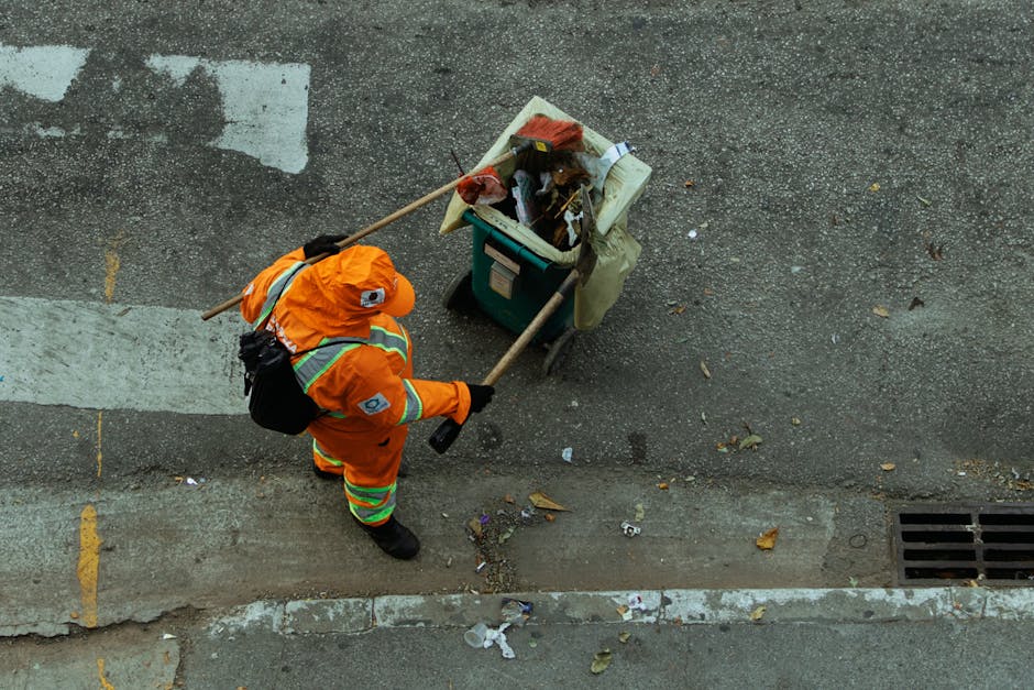 A street scene showing a woman in an orange reflective vest sweeping the pavement with a traditional broom made of straw. She is wearing a dark dress and a headscarf, with a face mask for protection.