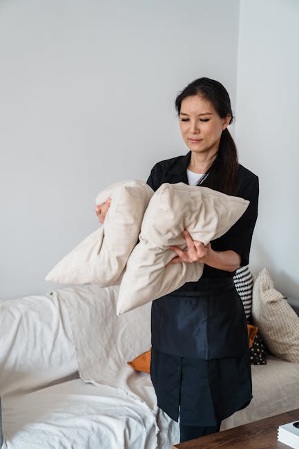 A person holding a vacuum cleaner with a flexible hose is performing surface cleaning on a decorative cushion with a yellow and white geometric pattern, placed on a dark fabric sofa in a well-lit room